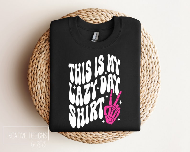 Puede incluir: Camiseta negra con texto blanco que dice "This is my lazy day shirt" en una fuente retro. Una mano de esqueleto rosa con un signo de paz est&aacute; a la derecha del texto.