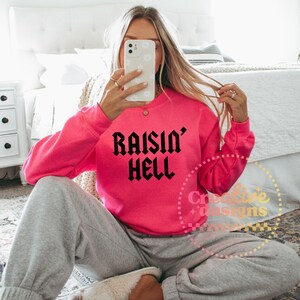 May include: A vibrant pink sweatshirt featuring the text "Raisin' Hell" in black gothic font. The top is paired with grey sweatpants. The image is taken indoors.