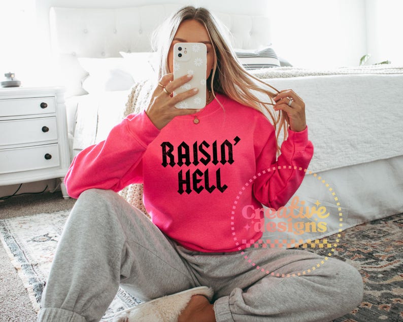 May include: A vibrant pink sweatshirt featuring the text "Raisin' Hell" in black gothic font. The top is paired with grey sweatpants. The image is taken indoors.