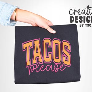 May include: A black t-shirt with the words "TACOS PLEASE" in orange and pink lettering. The text is in a collegiate style font. The words "creative designs by TSC" are written in a script font in the upper right corner.