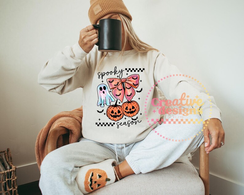 Puede incluir: Sudadera beige con un dise&ntilde;o de Halloween. El dise&ntilde;o incluye un fantasma, un lazo, dos calabazas y las palabras "spooky season". La sudadera se combina con pantalones de ch&aacute;ndal grises y un gorro color canela.