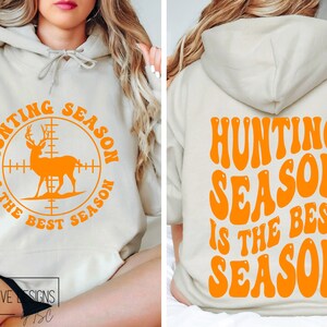 May include: A beige hoodie with orange text and graphics. The front features a deer silhouette inside a crosshair circle with the text "HUNTING SEASON IS THE BEST SEASON". The back of the hoodie repeats the phrase in a retro font.