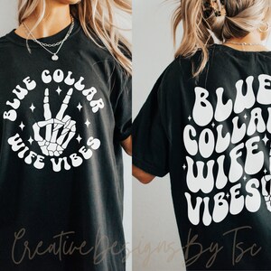 May include: Black t-shirt with white text that reads "Blue Collar Wife Vibes" in a retro font. A skeleton hand with a peace sign is in the center of the text.
