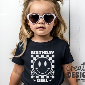 May include: A black short-sleeved t-shirt featuring a white graphic print of a smiley face making a peace sign, set against a checkered background. The text "BIRTHDAY GIRL" is printed in white above and below the graphic. White heart-shaped sunglasses.