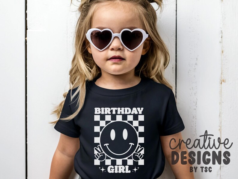 May include: A black short-sleeved t-shirt featuring a white graphic print of a smiley face making a peace sign, set against a checkered background. The text "BIRTHDAY GIRL" is printed in white above and below the graphic. White heart-shaped sunglasses.