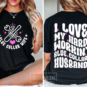 May include: Black t-shirt with a white graphic design featuring a wrench, screwdriver, diamond ring, and heart. The text reads "Blue Collar Wife". The back of the shirt has white text that reads "I Love My Hard Workin' Blue Collar Husband".