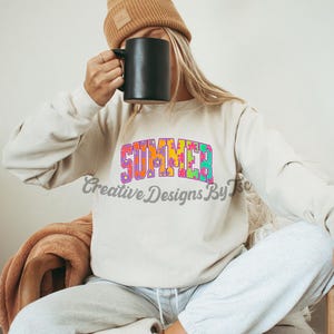 Puede incluir: Una sudadera blanca con la palabra "SUMMER" impresa en letras coloridas de tie-dye. La sudadera la lleva puesta una persona que sostiene una taza negra.