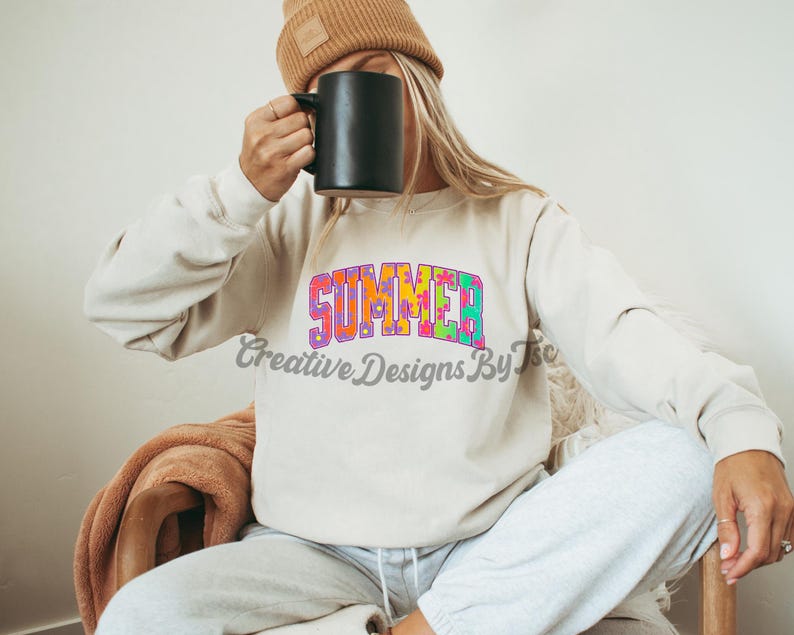 Puede incluir: Una sudadera blanca con la palabra "SUMMER" impresa en letras coloridas de tie-dye. La sudadera la lleva puesta una persona que sostiene una taza negra.