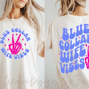 May include: A white t-shirt with the text "BLUE COLLAR WIFE VIBES" in blue and pink lettering with a pink skeleton hand making a peace sign in the center of the text. The text is in a retro font style.