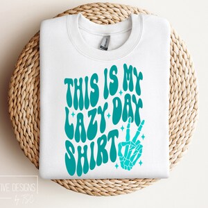 Puede incluir: Sudadera blanca con un gr&aacute;fico azul turquesa que dice "This is my lazy day shirt" con una mano de esqueleto haciendo un signo de paz.