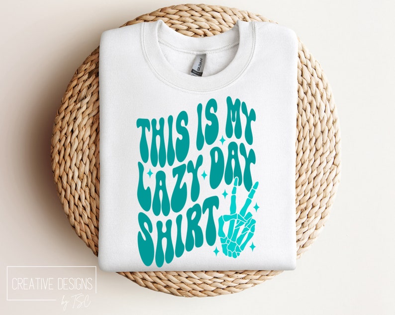 Puede incluir: Sudadera blanca con un gr&aacute;fico azul turquesa que dice "This is my lazy day shirt" con una mano de esqueleto haciendo un signo de paz.