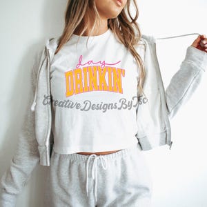 Op de afbeelding: Een wit crop top met de tekst "day drinkin'" in oranje en roze letters. Het topje wordt gedragen door een persoon met lang blond haar en wordt gecombineerd met een grijze joggingbroek.