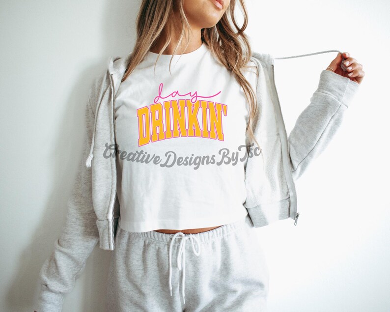 Op de afbeelding: Een wit crop top met de tekst "day drinkin'" in oranje en roze letters. Het topje wordt gedragen door een persoon met lang blond haar en wordt gecombineerd met een grijze joggingbroek.