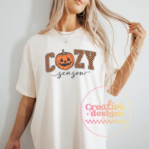 May include: Cream-colored t-shirt with the words "COZY season" in a fall-themed design. The word "COZY" is in orange and brown checkered letters, with an orange jack-o'-lantern in the center. The word "season" is written in a cursive font.