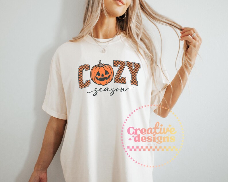 May include: Cream-colored t-shirt with the words "COZY season" in a fall-themed design. The word "COZY" is in orange and brown checkered letters, with an orange jack-o'-lantern in the center. The word "season" is written in a cursive font.