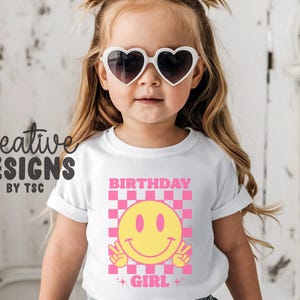 May include: A white t-shirt featuring a graphic print with a pink and white checkered square background. Centered is a yellow smiley face making a peace sign with both hands. The text BIRTHDAY GIRL is printed in pink above and below the graphic.