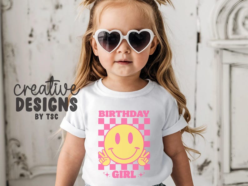 May include: A white t-shirt featuring a graphic print with a pink and white checkered square background. Centered is a yellow smiley face making a peace sign with both hands. The text BIRTHDAY GIRL is printed in pink above and below the graphic.