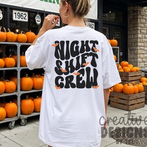 May include: A white short-sleeved t-shirt featuring the black text NIGHT SHIFT CREW in a wavy, retro-inspired font on the back. The design is accented with small, scattered orange bat illustrations throughout the lettering.