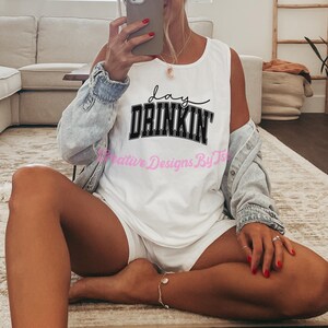 Op de afbeelding: Een wit tanktop met zwarte tekst die "Lay Drinkin'" zegt. Het tanktop wordt gedragen door een persoon die op een lichtbruin tapijt zit.