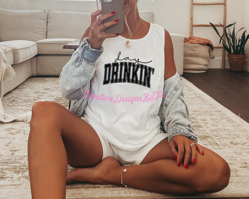 Op de afbeelding: Een wit tanktop met zwarte tekst die "Lay Drinkin'" zegt. Het tanktop wordt gedragen door een persoon die op een lichtbruin tapijt zit.