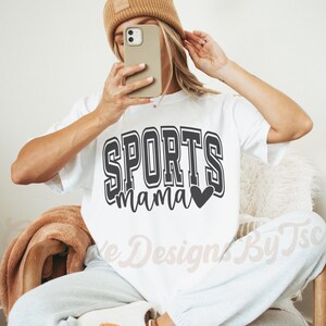 May include: White t-shirt with black text that reads "SPORTS mama" with a small black heart. The shirt is being worn by a person with a brown beanie.
