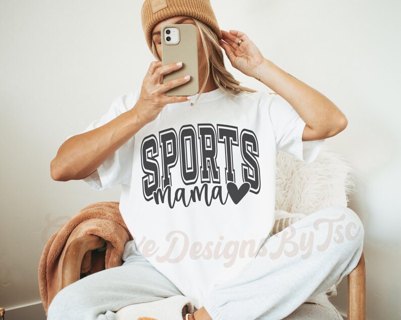 May include: White t-shirt with black text that reads "SPORTS mama" with a small black heart. The shirt is being worn by a person with a brown beanie.