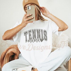 May include: White t-shirt with the text "Tennis Mama" in gray with a small heart graphic. The model is wearing the shirt and has a brown beanie on.