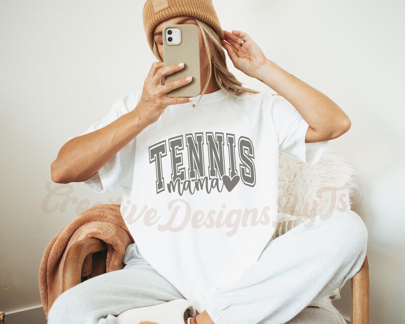 May include: White t-shirt with the text "Tennis Mama" in gray with a small heart graphic. The model is wearing the shirt and has a brown beanie on.