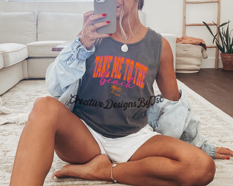 K&ouml;nnte beinhalten: Ein graues Tanktop mit dem Text "Take Me To The Beach" in orangefarbenen und rosafarbenen Buchstaben. Das Oberteil zeigt ein Palmenmotiv und wird von einer Person mit einem wei&szlig;en Rock getragen.