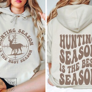 May include: Beige hoodie with the text "HUNTING SEASON IS THE BEST SEASON" on the front and back. The front design features a deer inside a crosshair circle. The back design has the same text in a retro font.