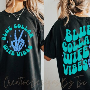 May include: Black t-shirt with a blue and teal graphic design that says "Blue Collar Wife Vibes" with a peace sign skeleton hand.
