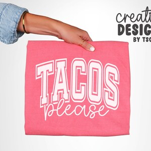 May include: A folded pink t-shirt with the words "TACOS PLEASE" in white block letters. The words are in a collegiate style font. The shirt is held by a person wearing a denim jacket.
