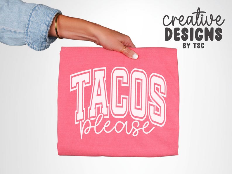 May include: A folded pink t-shirt with the words "TACOS PLEASE" in white block letters. The words are in a collegiate style font. The shirt is held by a person wearing a denim jacket.