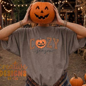 May include: A grey short-sleeved t-shirt featuring a graphic print with the text "COZY season" in a black and orange checkered font. The design includes a central orange jack-o'-lantern illustration. The shirt is made of cotton fabric.