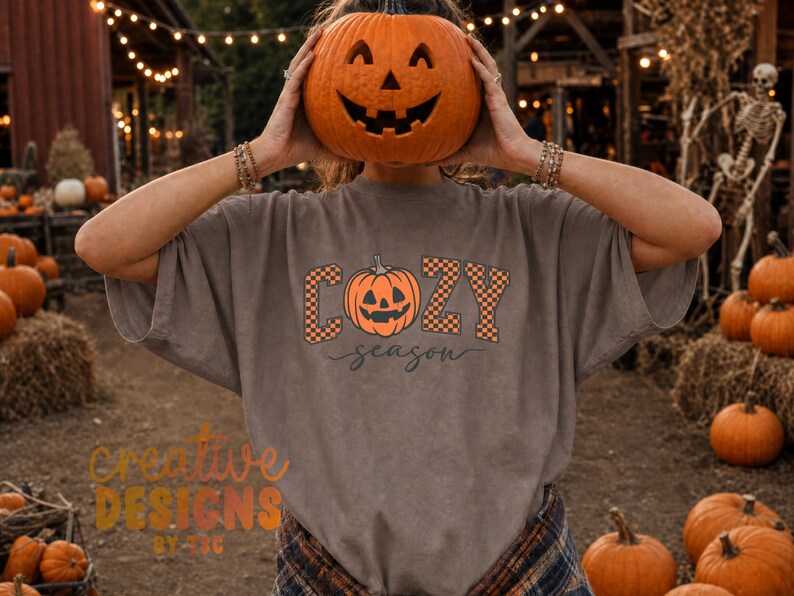 May include: A grey short-sleeved t-shirt featuring a graphic print with the text "COZY season" in a black and orange checkered font. The design includes a central orange jack-o'-lantern illustration. The shirt is made of cotton fabric.