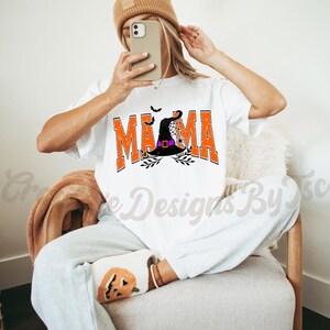 Puede incluir: Camiseta blanca con un gr&aacute;fico naranja y negro que dice "Mama" con un sombrero de bruja y un dise&ntilde;o de telara&ntilde;a.