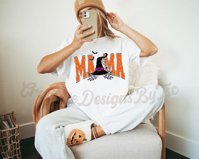 Puede incluir: Camiseta blanca con un gr&aacute;fico naranja y negro que dice "Mama" con un sombrero de bruja y un dise&ntilde;o de telara&ntilde;a.