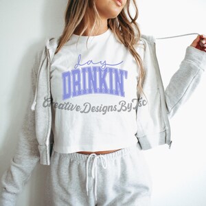 Op de afbeelding: Een wit crop top met de tekst "day drinkin'" in paarse letters. Het top wordt gedragen met een grijze hoodie en grijze sweatpants.