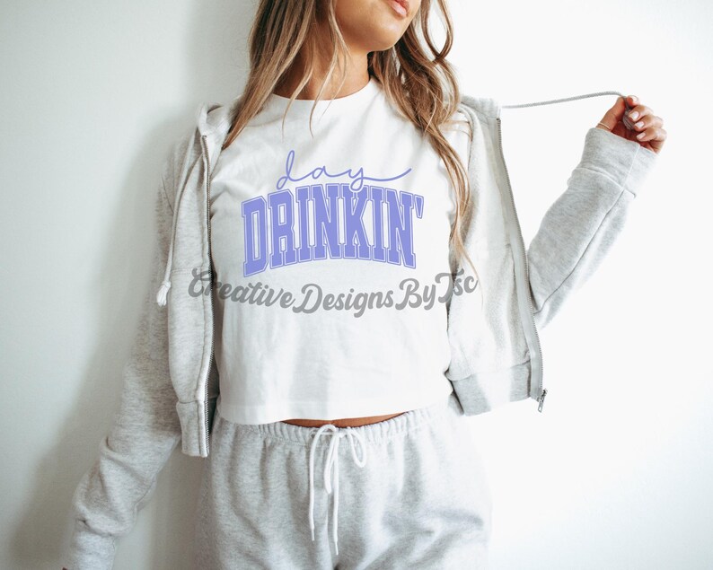 Op de afbeelding: Een wit crop top met de tekst "day drinkin'" in paarse letters. Het top wordt gedragen met een grijze hoodie en grijze sweatpants.