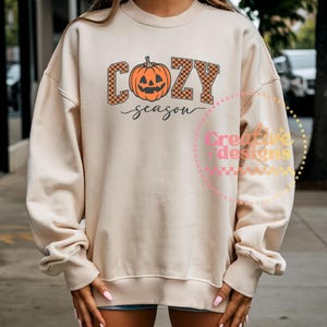 May include: A cream-colored sweatshirt with the words "COZY season" in orange and black checkered letters and a pumpkin graphic. The sweatshirt has long sleeves and a crew neck.