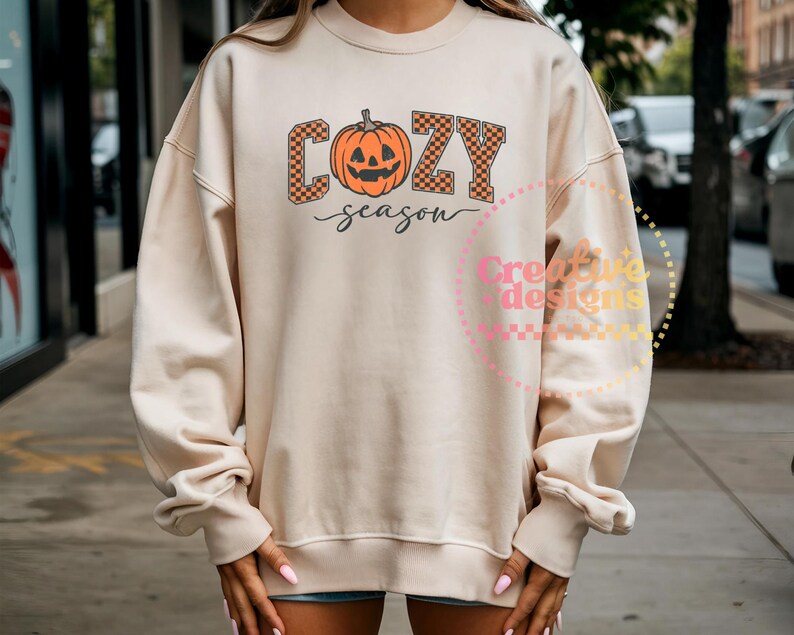 May include: A cream-colored sweatshirt with the words "COZY season" in orange and black checkered letters and a pumpkin graphic. The sweatshirt has long sleeves and a crew neck.