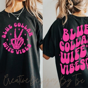 May include: Black t-shirt with a pink graphic that says "Blue Collar Wife Vibes" with a skeleton hand making a peace sign.