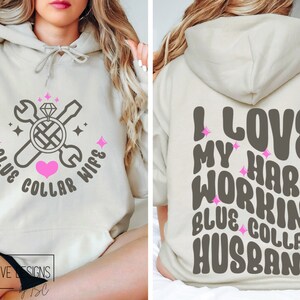 May include: A light beige hoodie with a graphic design on the front and back. The front design features a wrench, screwdriver, and a diamond ring with the text "Blue Collar Wife". The back design features the text "I Love My Hard Workin' Blue Collar Husband".