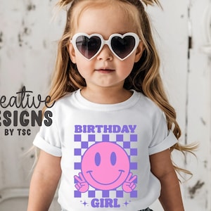 May include: A white short-sleeved t-shirt featuring a graphic print of a pink smiley face making peace signs, set against a purple and white checkered background. The text "BIRTHDAY GIRL" is printed in purple above and below the graphic.