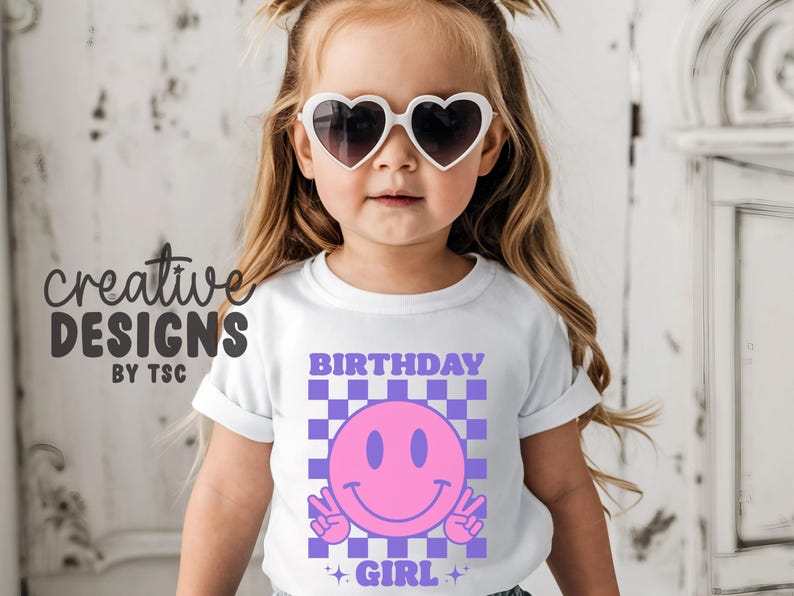 May include: A white short-sleeved t-shirt featuring a graphic print of a pink smiley face making peace signs, set against a purple and white checkered background. The text "BIRTHDAY GIRL" is printed in purple above and below the graphic.
