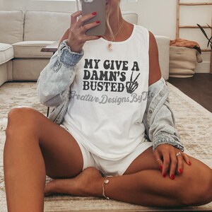 May include: White tank top with black text that reads "My Give A Damn's Busted" with a peace sign and a hand graphic. The tank top is being worn by a person sitting on a rug.