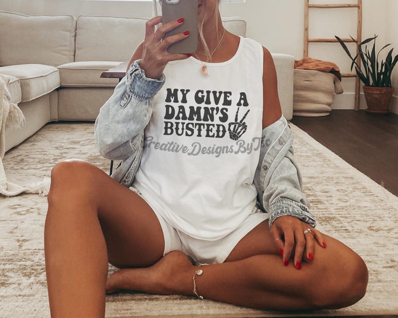 May include: White tank top with black text that reads "My Give A Damn's Busted" with a peace sign and a hand graphic. The tank top is being worn by a person sitting on a rug.