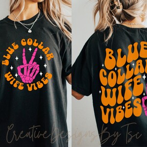 May include: Black t-shirt with an orange graphic that says "Blue Collar Wife Vibes" with a pink skeleton hand making a peace sign.