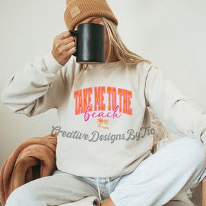 K&ouml;nnte beinhalten: Ein wei&szlig;er Pullover mit dem Text "Take Me To The Beach" in rosa und orangefarbenen Buchstaben. Der Text ist von Palmen und einer Sonne umgeben.