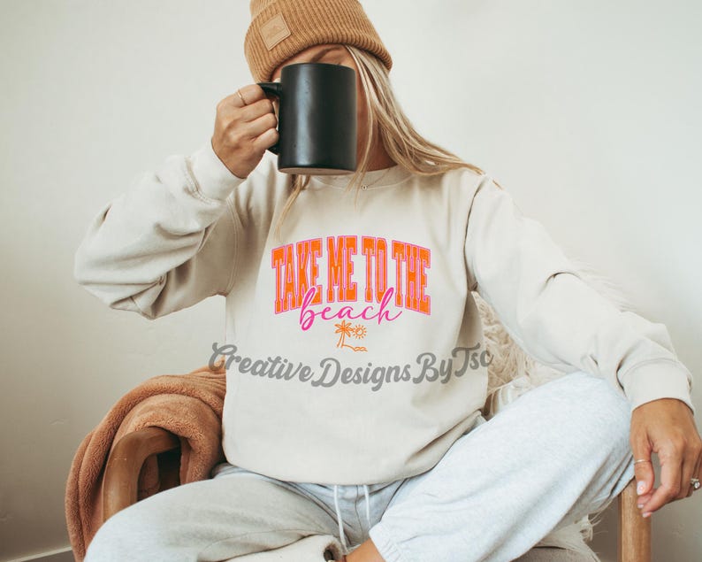 K&ouml;nnte beinhalten: Ein wei&szlig;er Pullover mit dem Text "Take Me To The Beach" in rosa und orangefarbenen Buchstaben. Der Text ist von Palmen und einer Sonne umgeben.